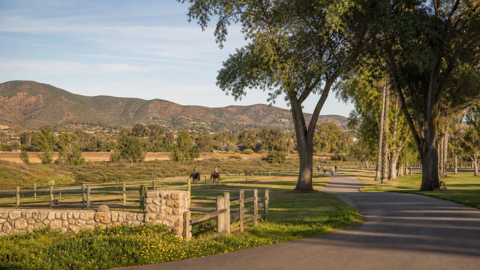 A paved path beside a wooden ranch fence, horses grazing in the field beyond and hills behind