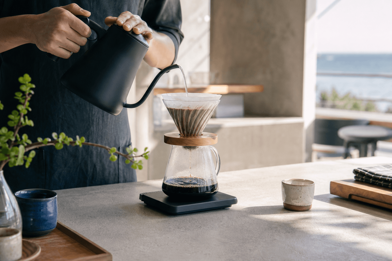A barista pours water from a black gooseneck kettle over a coffee dripper in morning light