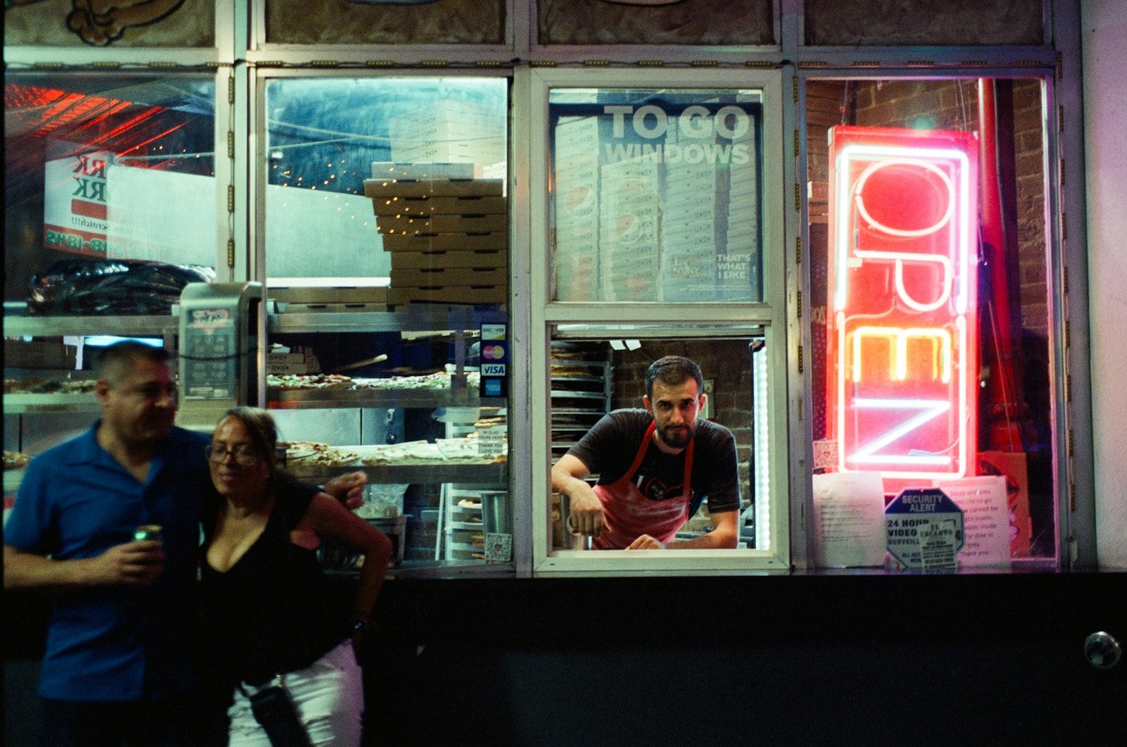A lit to-go window at a late-night pizza counter, stacked boxes and a neon open sign