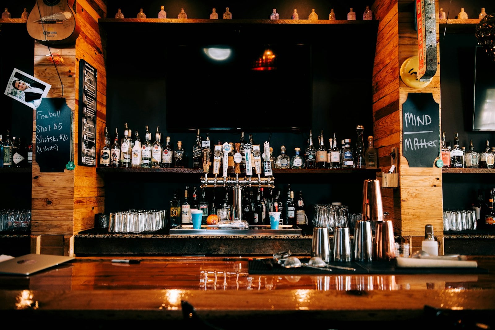 A worn wooden bar counter with taps, a full back shelf and a chalkboard drink list