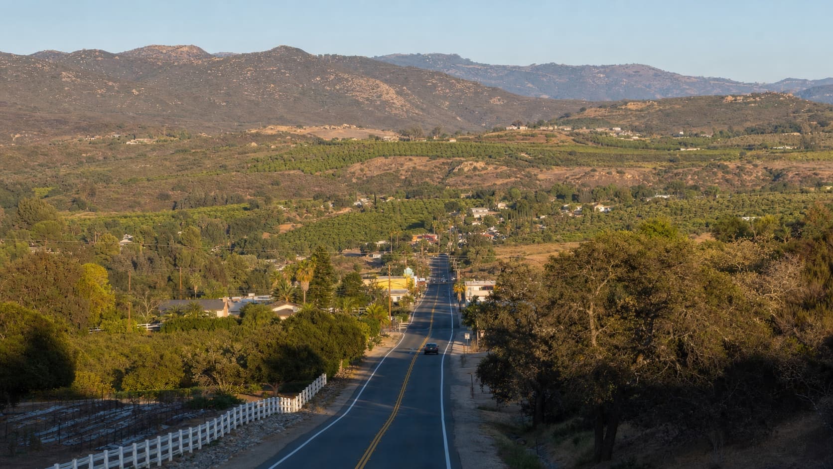 A two-lane highway dropping into a valley of orchards and groves with bare mountains behind