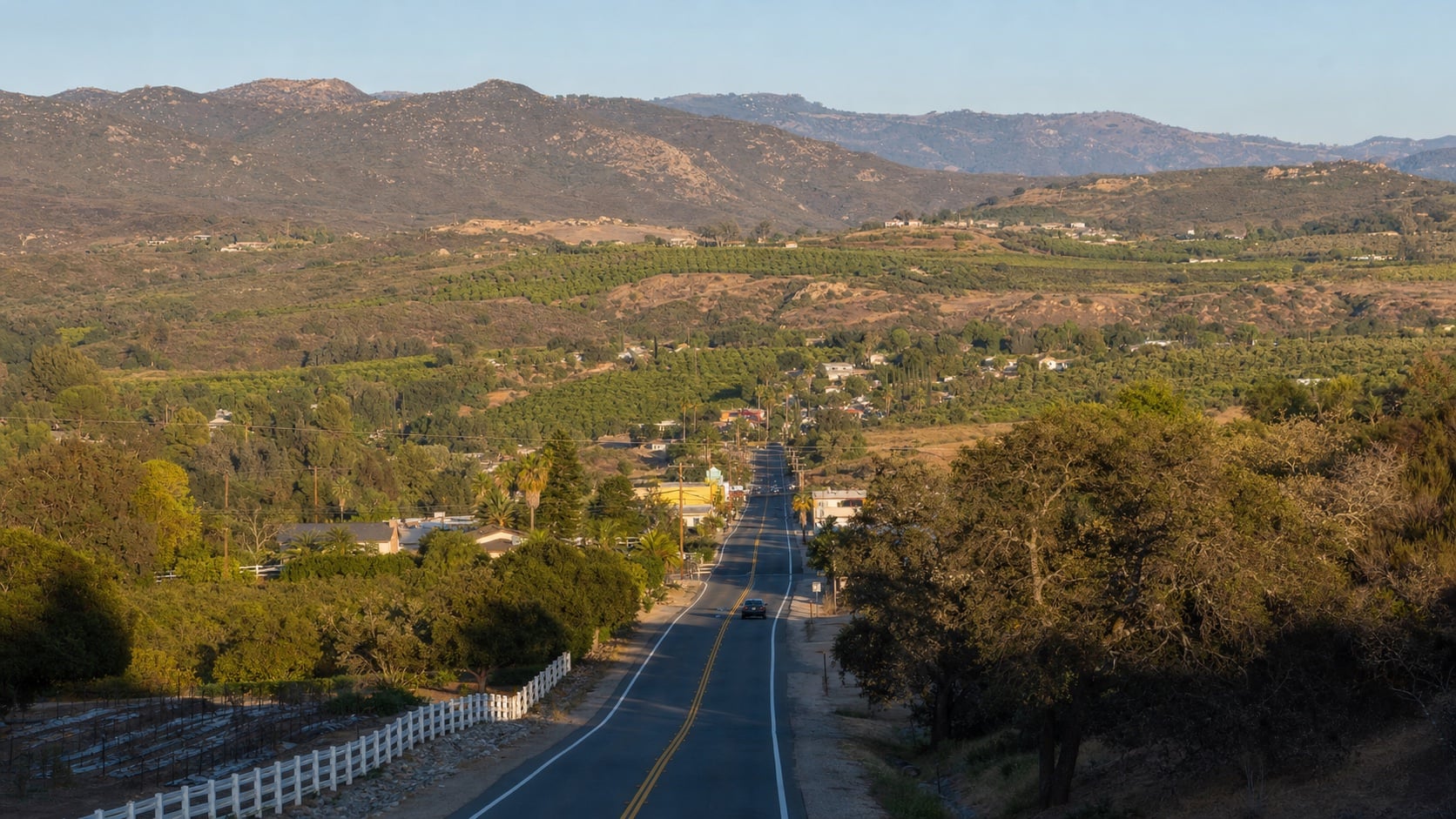 Rural inland North County road running through green groves toward dry foothills