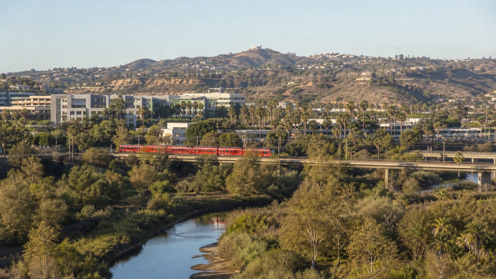 A red San Diego Trolley crossing the green San Diego River corridor in Mission Valley