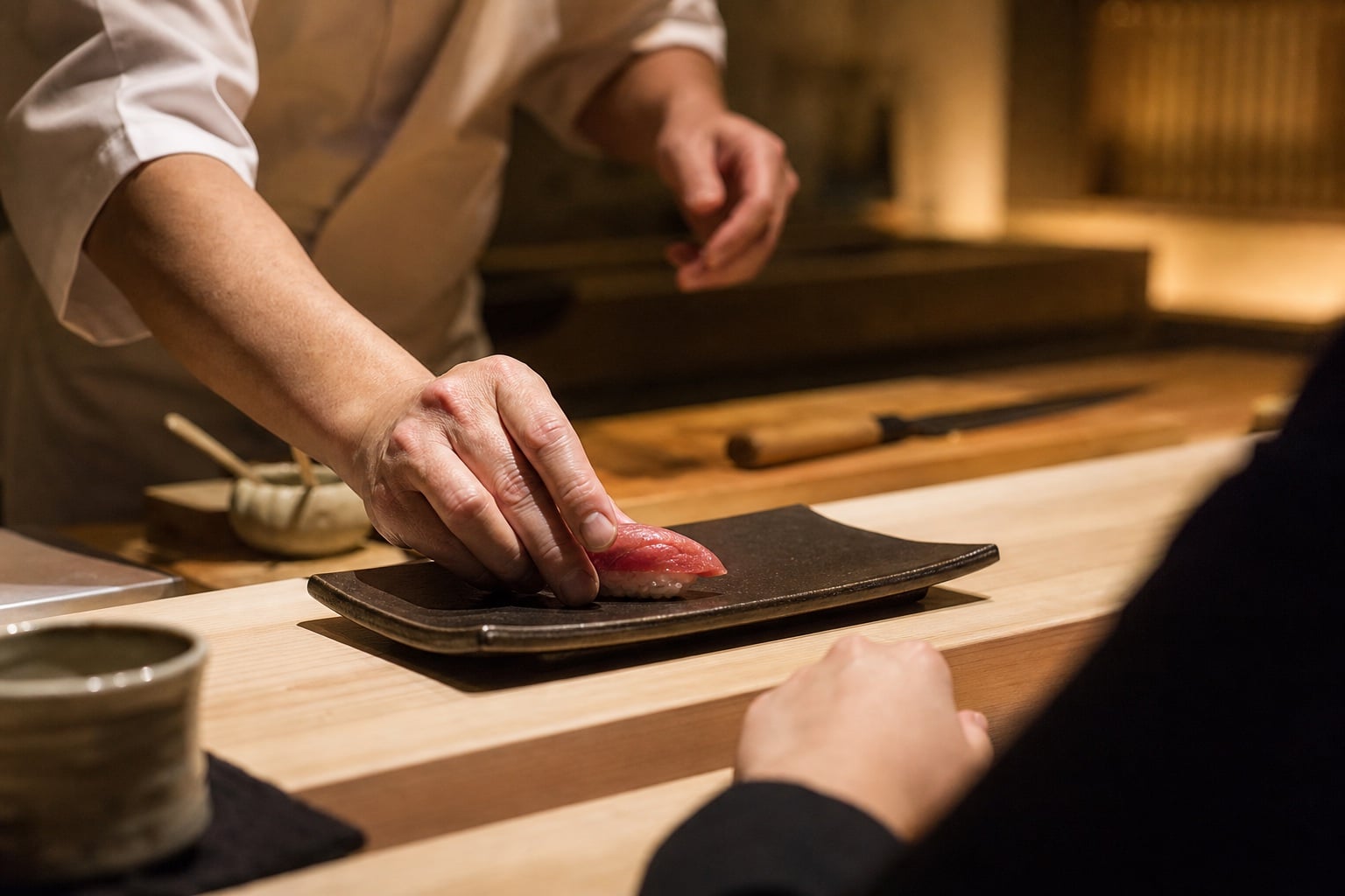 Sushi chef placing tuna nigiri on a dark plate at a wood counter