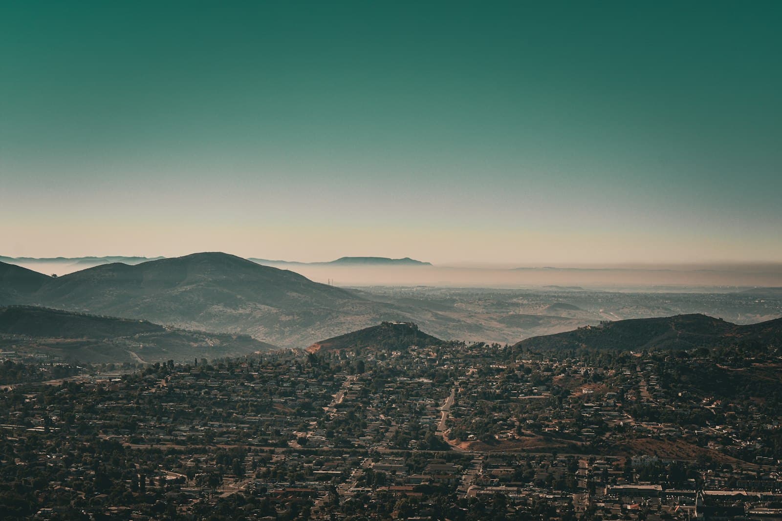 A hazy valley of low hills with a town spread across the floor below, under a teal sky