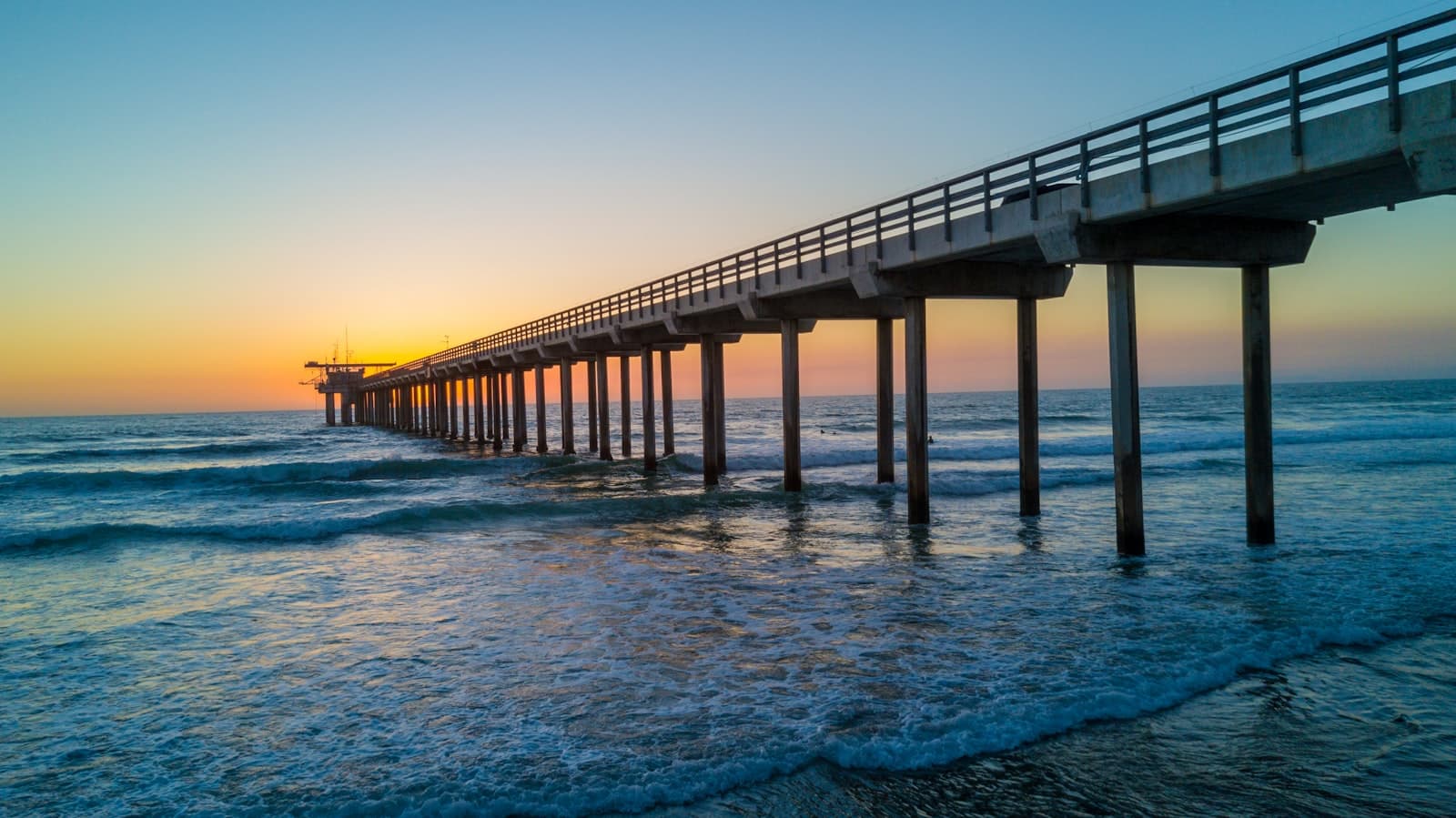 A long pier on slender pilings reaching out over breaking surf at sunset