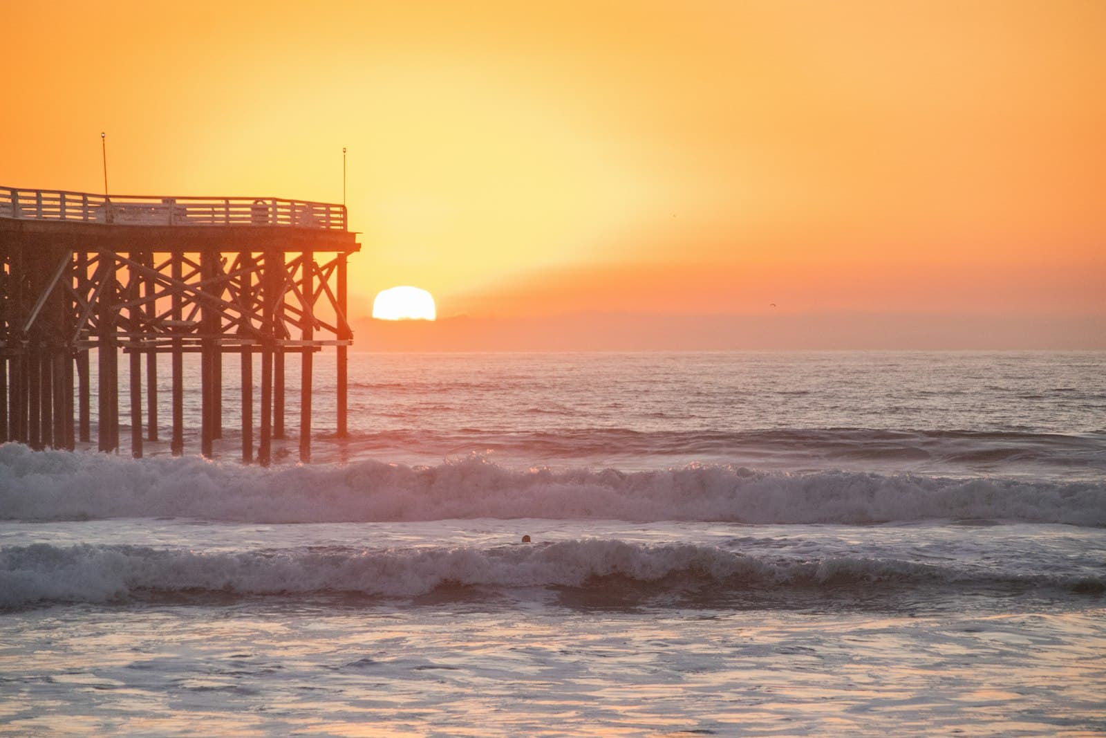The sun dropping into the ocean beside the silhouetted pier, waves rolling in below