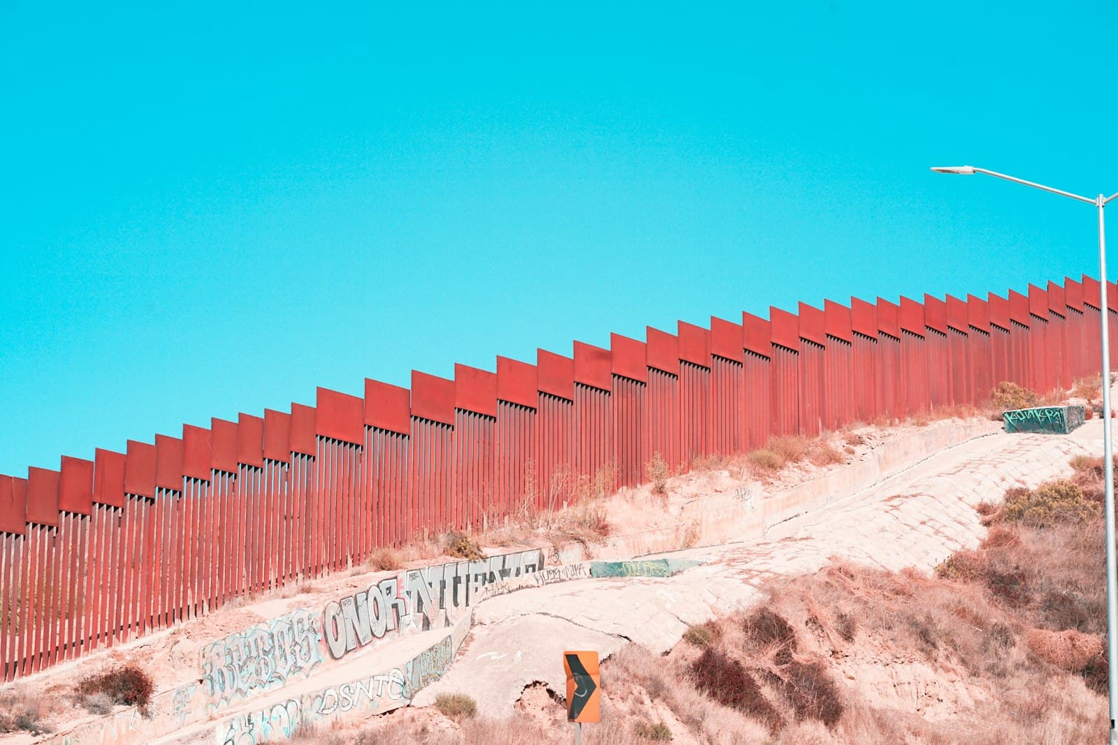 A tall rust-red border fence running along a hillside against a turquoise sky